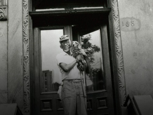 Black and white photo featuring an elderly man holding a dog in the doorway of a brownstone apartment building