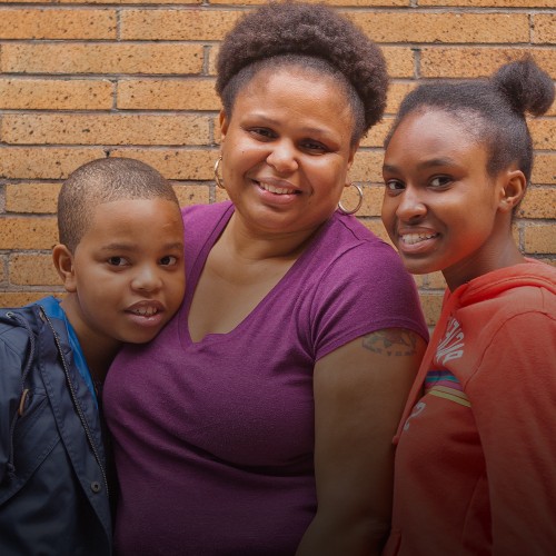 A male child in a blue jacket, a woman in a purple t-shirt, and a female child in an orange hoodie pose in front of a brick wall.