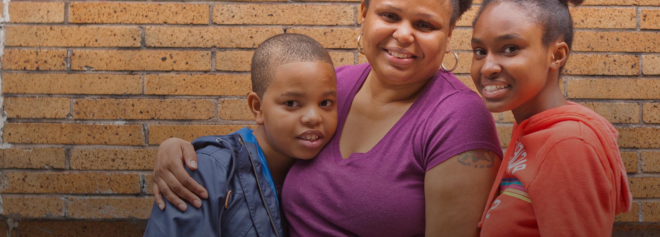 A male child in a blue jacket, a woman in a purple t-shirt, and a female child in an orange hoodie pose in front of a brick wall.