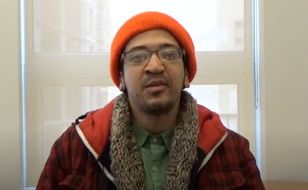 An adult with glasses, an orange hat, a plaid sweater, a brown scarf, and a green shirt underneath. The person is sitting down looking directly at the camera.