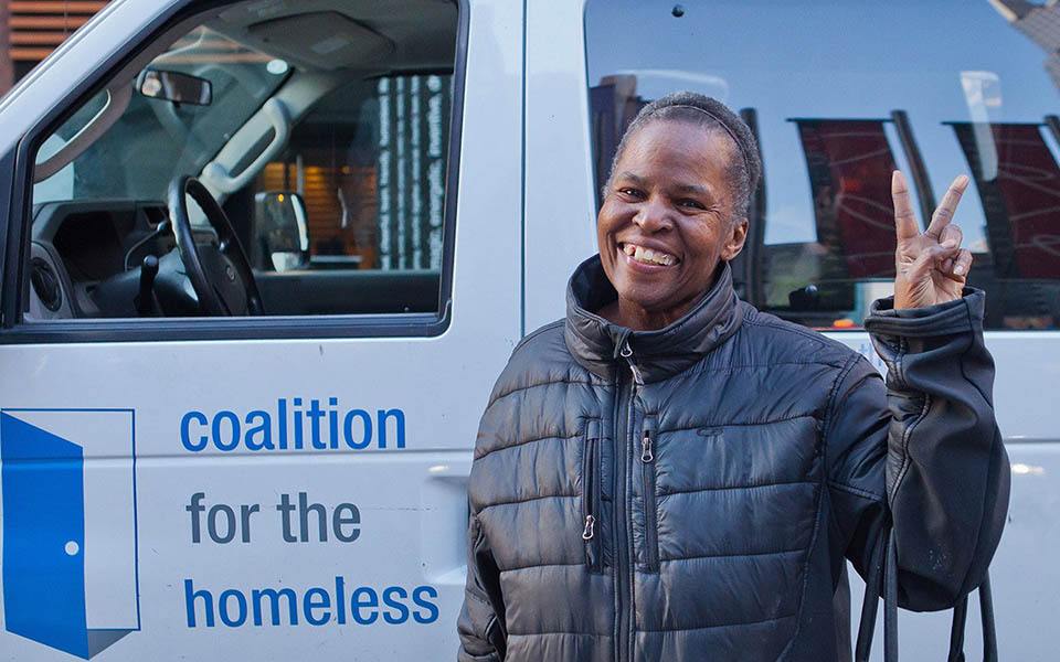A woman in a coat holds up a peace sign in front of a Coalition for the Homeless food van.