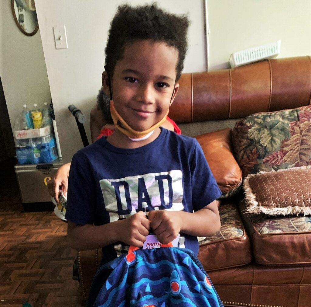 A boy smiles while holding up a blue backpack in his living room