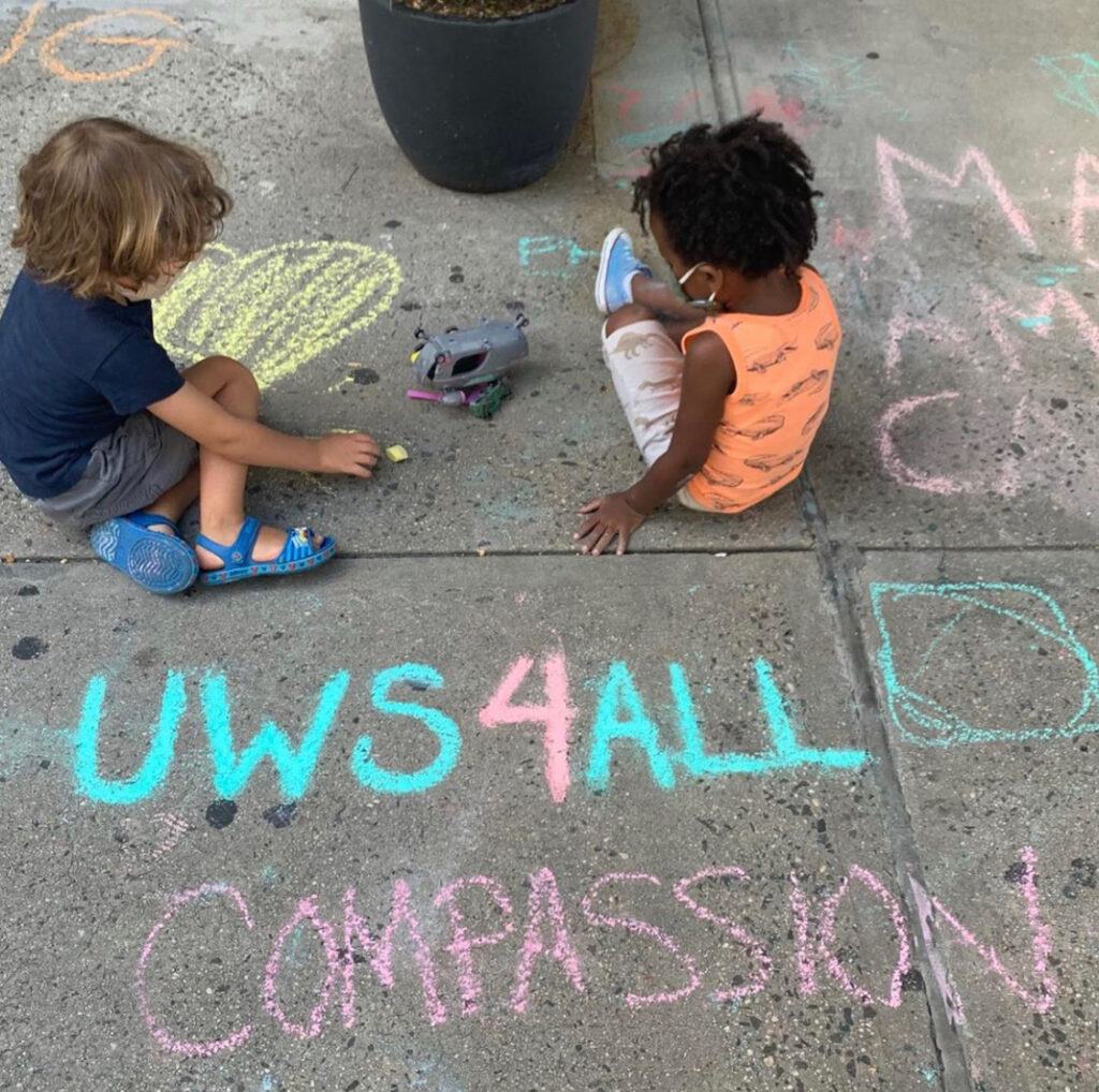 Two kids sitting on pavement surrounded by chalk drawings, the words "UWS4ALL" and "CoMPASSION" are written by them