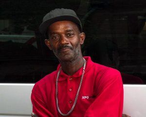 A man in a red-collared T-shirt with a blue baseball cap and a sliver chain leans against the side of the Coalition van, smiling softly at the camera with his arms crossed.