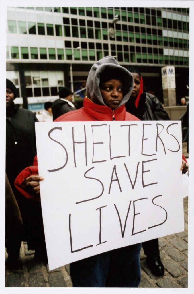 A sepia-toned photograph of a person in a red puffy winter jacket holding a large sign that says "Shelters Save Lives."