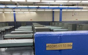 An off-white and blue room with florescent lights, blue poles, lockers, and beds with no sheets. In the forefront is a short blue wall with a brass sign that reads, "Rooms 177 to 196"
