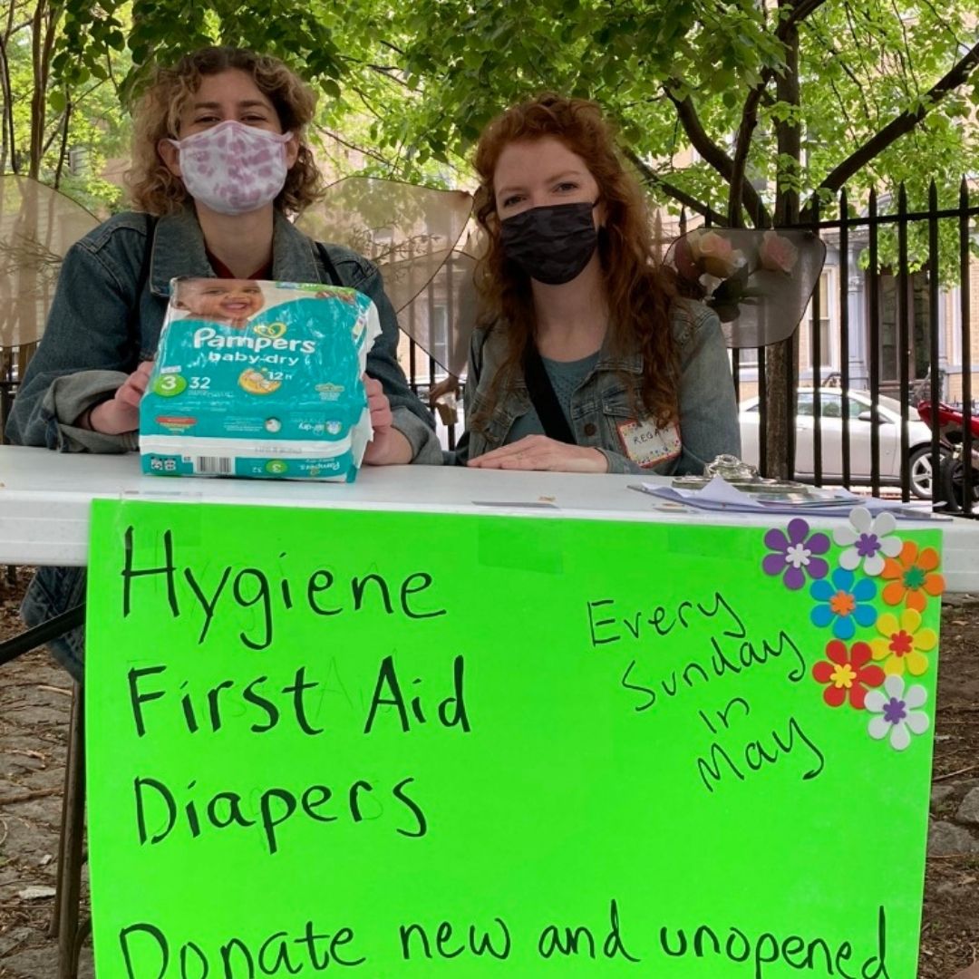 Two masked adults are sitting in a park behind a table holding up a package of Pampers. A lime green sign is taped to the table, reading "Hygiene, First Aid, Diapers, Donate new and unopened. Every Sunday in May. There are paper flowers on the top right corner of the sign.