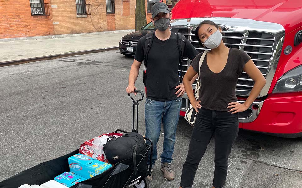 Two masked adults are standing in front of a red diesel truck holding a black wagon filled with packaged food, drinks, and other items
