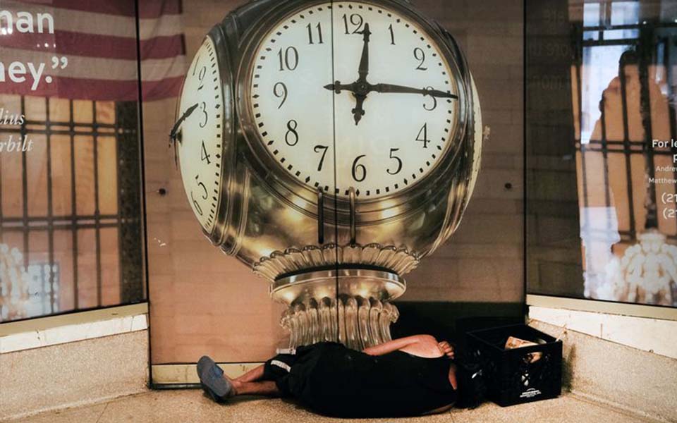 Individual wearing dark clothing is sleeping under a three dimensional clock. A gate is in the background.