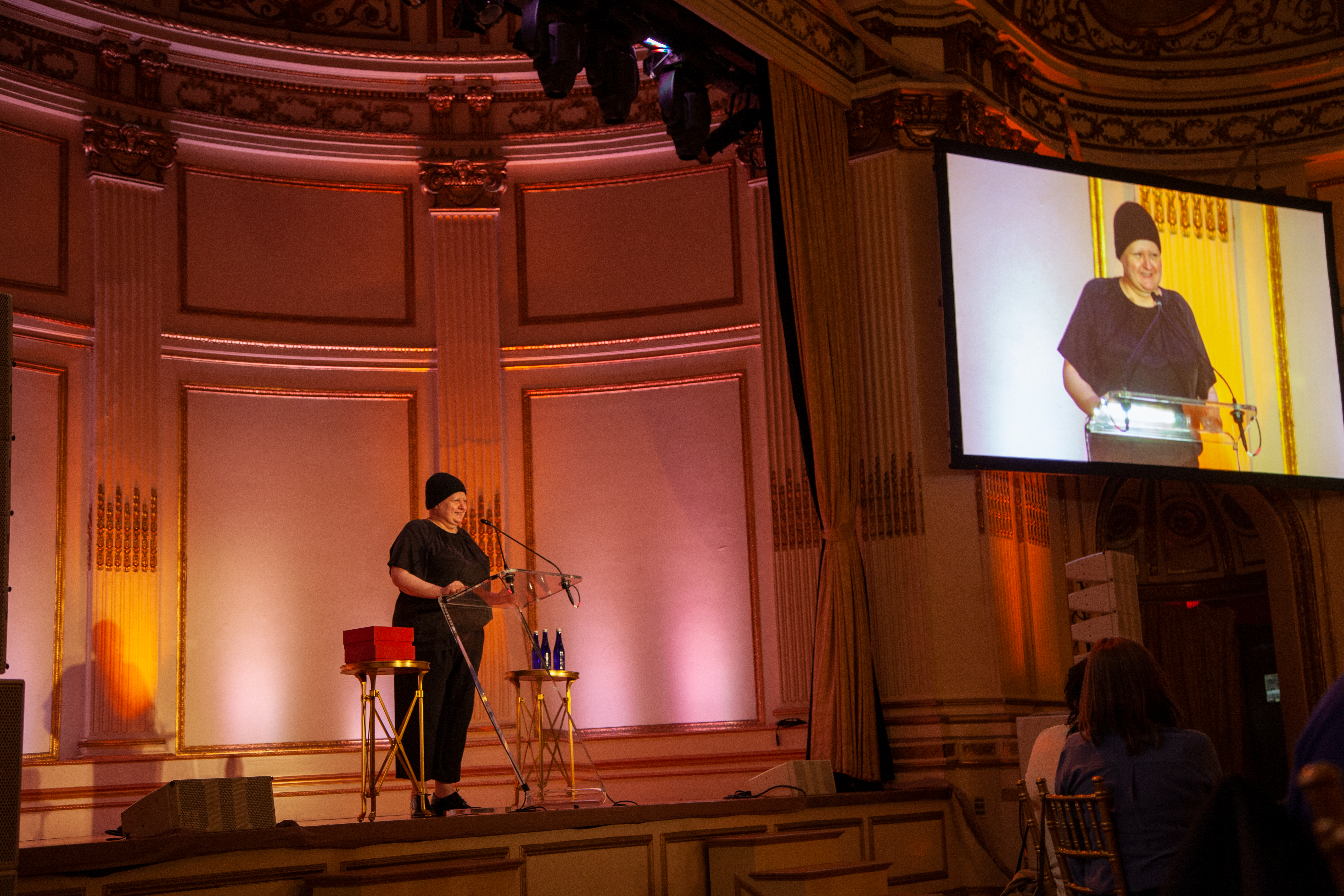Alexandra Waldman, Co-Founder and Creative Director of Universal Standard, wears all black with a black beanie and stands at a clear podium on a stage at the Plaza's Grand Ballroom.