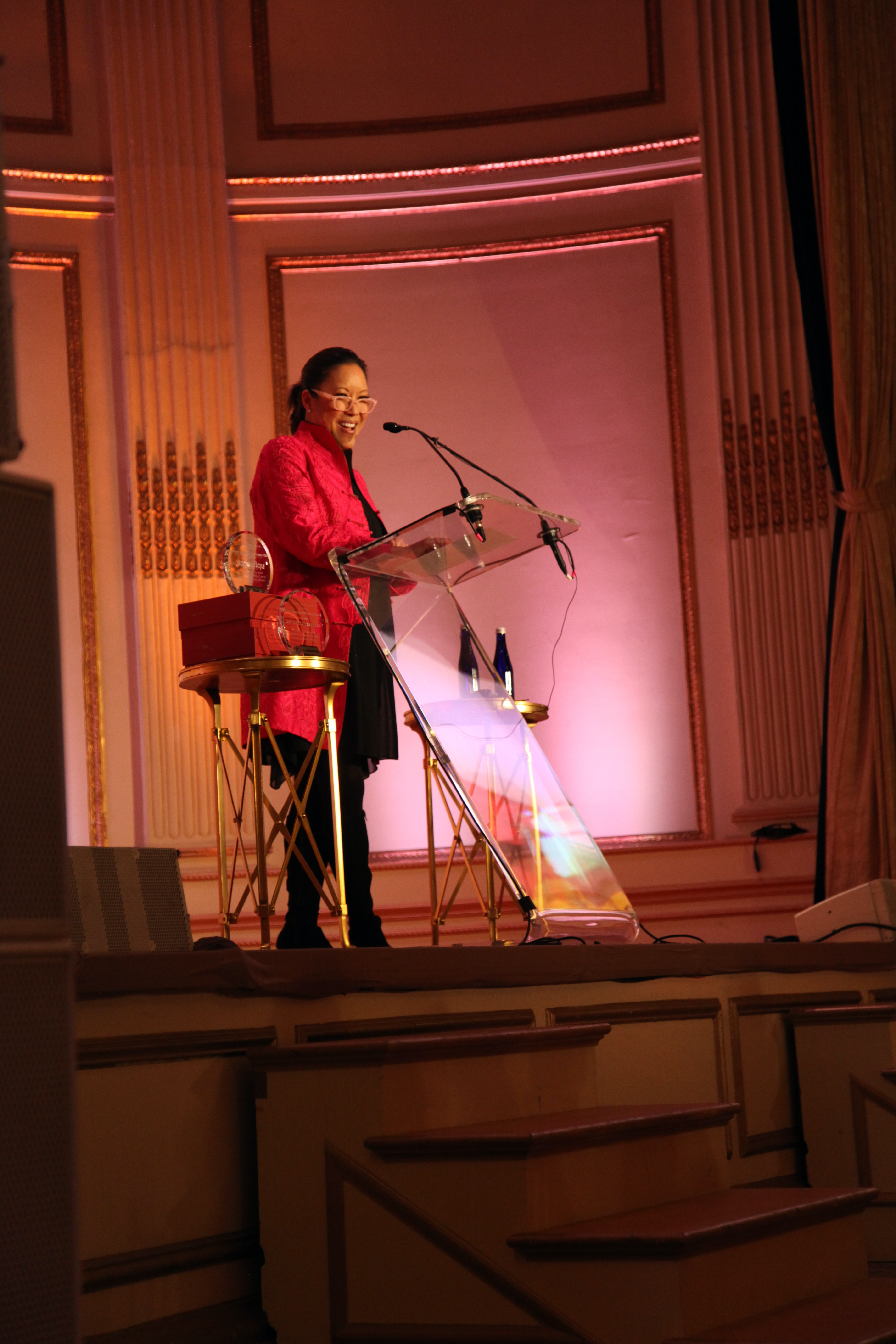 Emcee Cindy Hsu wears a bright pink blazer and stands at a clear podium on a stage in the Plaza's Grand Ballroom.