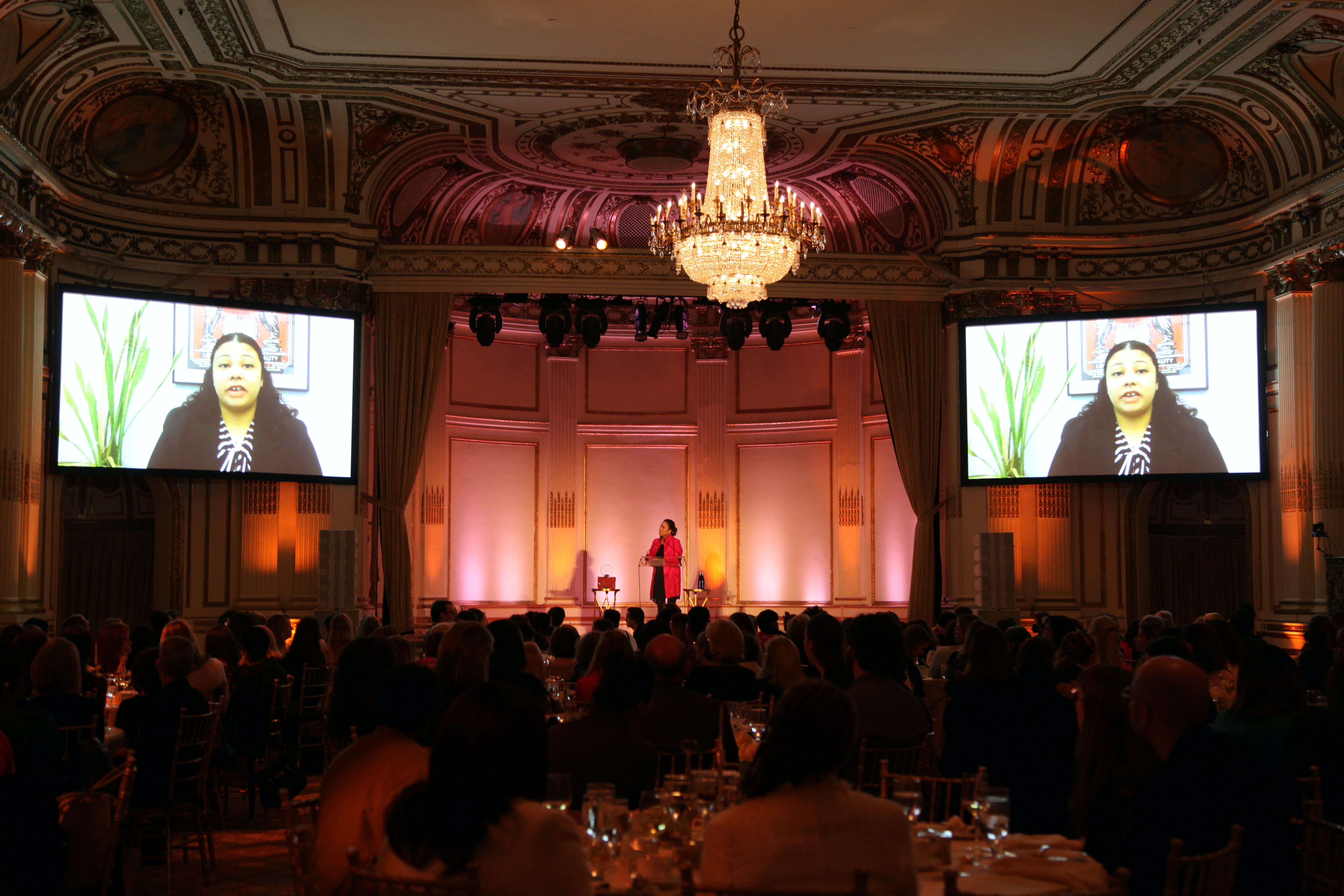 A large ballroom full of people sitting at round tables. In the front, two large screens on either side of a stage show the Zoom video of graduate speaker Paulette.