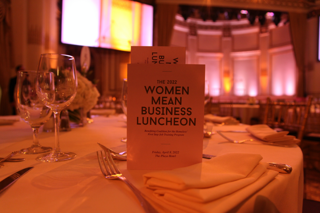 A program that reads, "The 2022 Women Mean Business Luncheon" stands upright on a table in a grand ballroom with a large stage and screen in the background.