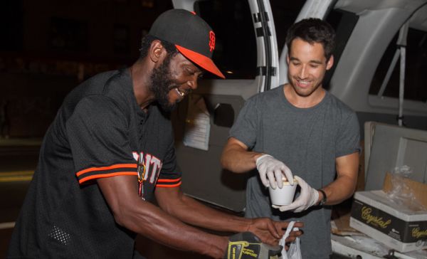 Two adults, one adult has a beard, wearing a black jersey with orange strips on the arms, and a black and orange hat, with one glove on his right hand. The other adult is wearing a gray t-shirt, and white latex gloves holding a white foam cup.