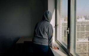 An adult by the window in an apartment wearing a gray sweater.