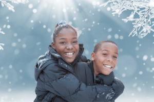 Two children wearing winter clothes and dusted with snow stand in front of a wintery scene. One is giving the other a hug.