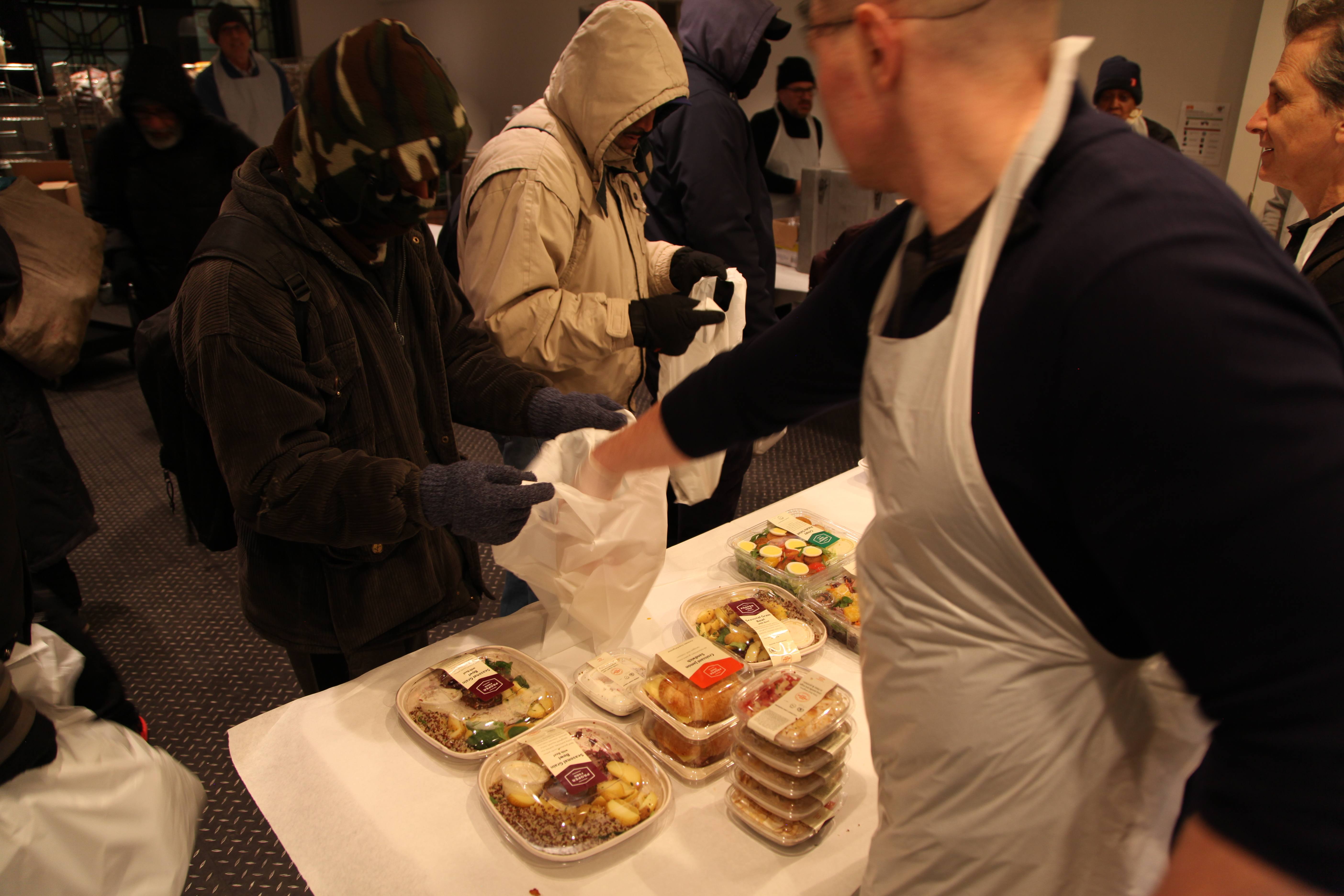Indoors, someone placing food into a bag that someone is holding out. In the foreground is a series of different meal options.