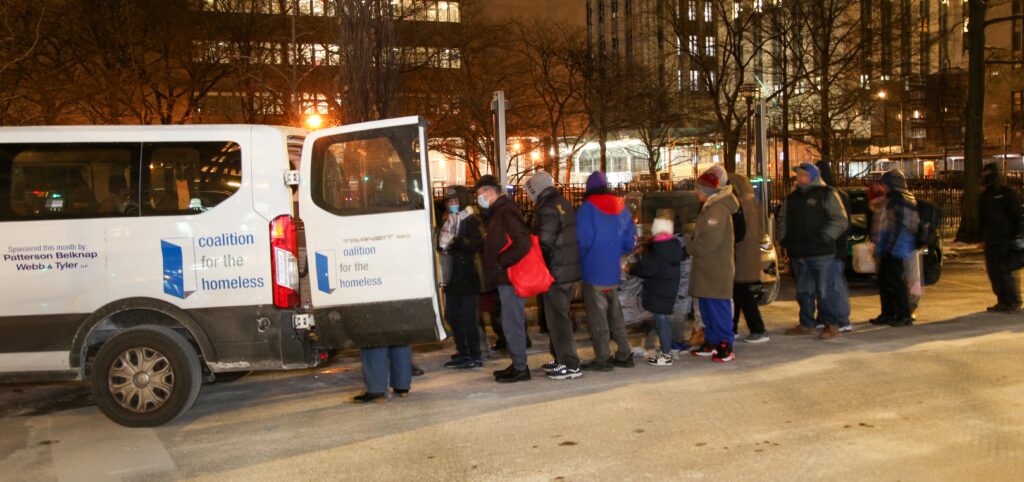 A Coalition for the Homeless van that has its barn doors open. There is a line of people, behind the van dressed in winters clothes. It is night time.