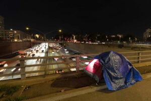 A lone tent on an overpass bridge while traffic goes back and forth below. It is night time.
