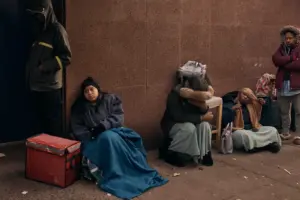 A line of sorrowful looking people, waiting on the street in winter clothes.
