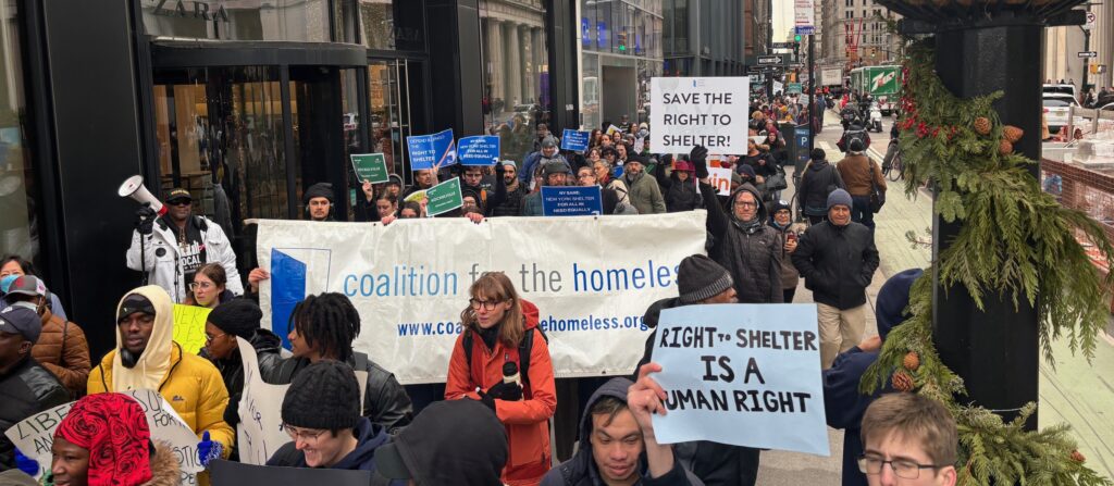 A group of protesters march down the street and are holding a sign that reads "Coalition for the Homeless" and "Save the Right to Shelter".