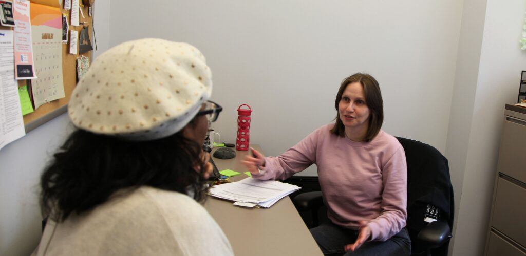 A women in the background is speaking facing towards someone in the foreground and having a conversation. They are sitting over a desk.