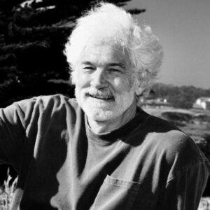 A black and white image of an older gentleman, smiling to someone off camera. The scene behind is countryside.