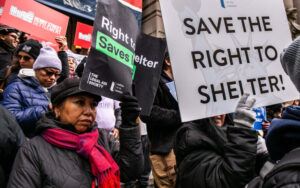 A group of people holding up signs at a protest. The signs read "save the right to shelter" and "right to shelter saves lives".