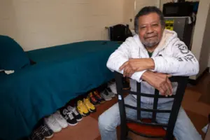 An image of a man sitting on a chair in front of a single bed, shoes alined down below it.