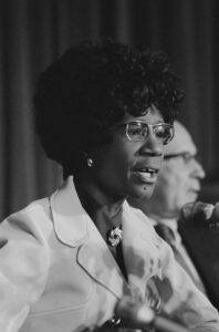 A black and white image of a woman (Shirley Chisholm) speaking into a microphone.