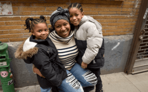 A parent embraces their young children. The young on the left wears a black jacket, their hair in braided pigtails. The old on the right, wears a winter jacket and has their hair in rows.