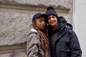 A couple stand close to each other in front of a sandstone wall. They are wearing winter coats, one has her hair in locks and dons a cap while the other wears a beanie. Their faces are somber, and expressions neutral. Image credit: Seze Devres