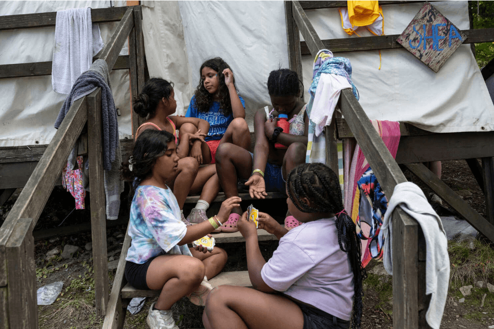 A group of six kids sitting on a a set of stairs in front of a canvas tent, playing a card game.