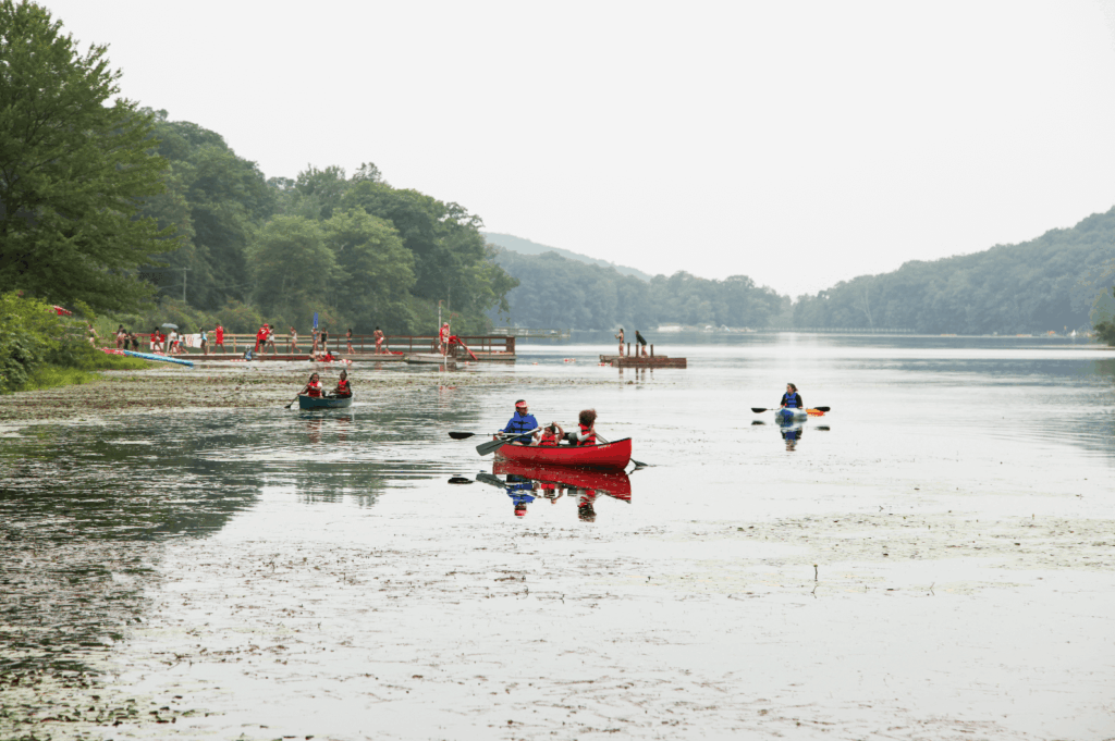 A still lake, dotted with canoes. The sky behind is cloudy, and they are surrounded by lush greenery.