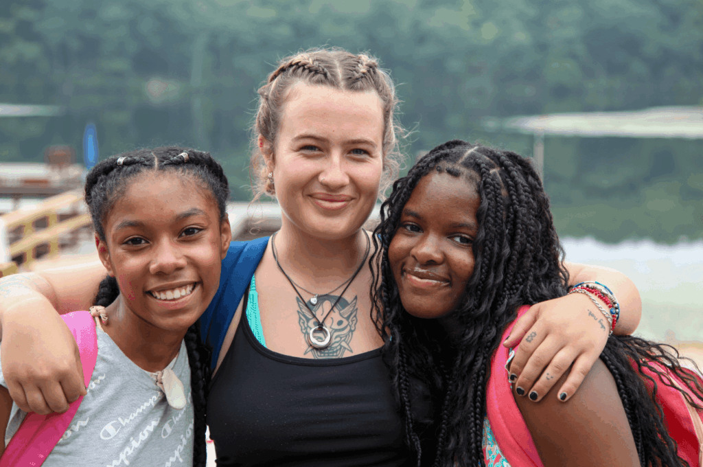 An older person has their arms around two teens. The teens where pink backpacks and all three smile gently towards the camera.