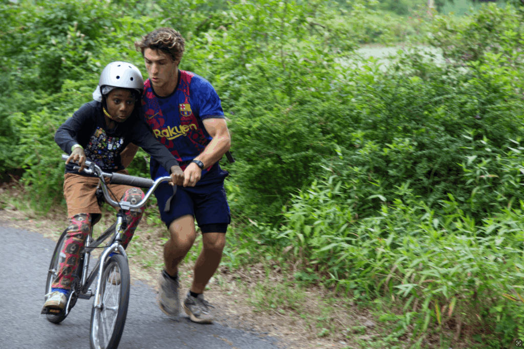 A young child is learning to ride a back, with the help of an adult, who is holding the bike steady. They are moving towards the camera, with lush greenery behind them.