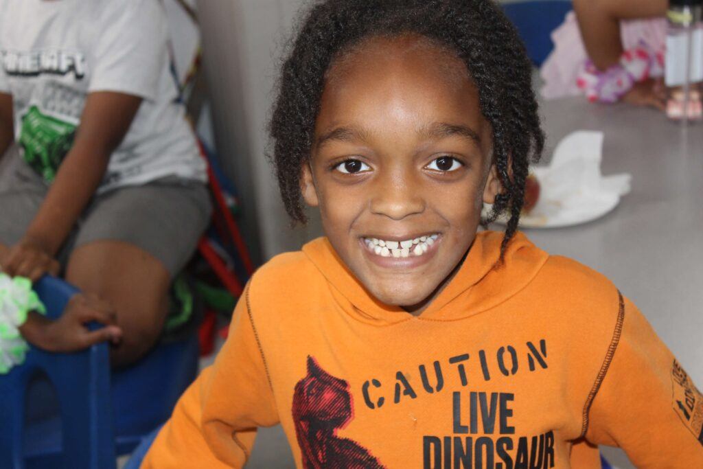 A kid, wearing an orange hoodie, smiles at the camera.