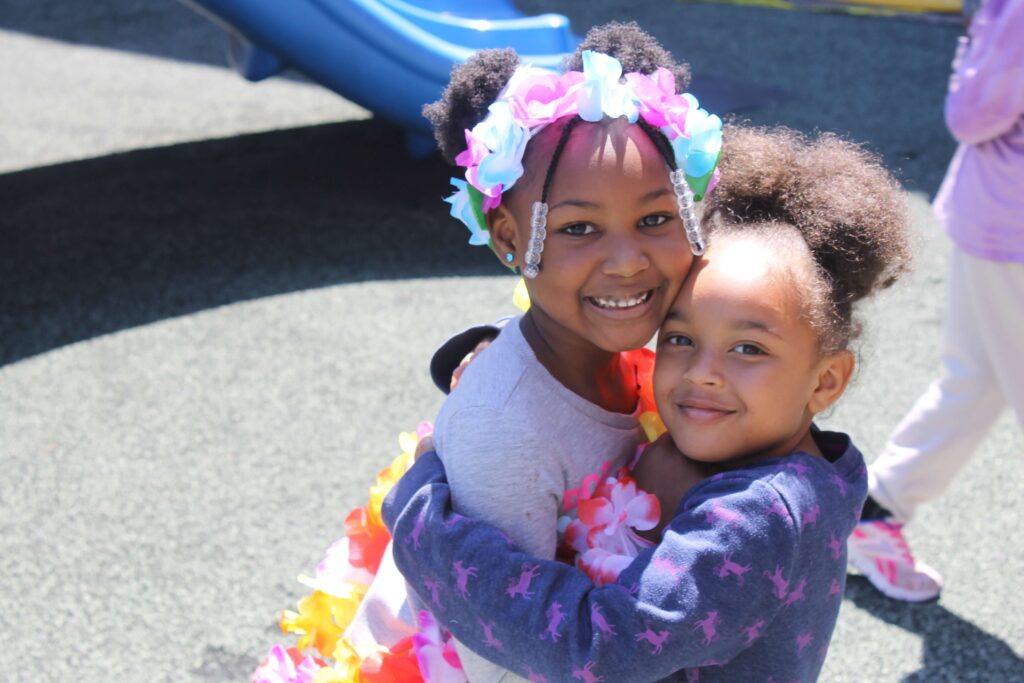 Two friends embrace, one wears a flower lei in their hair. The smile and look towards the camera.