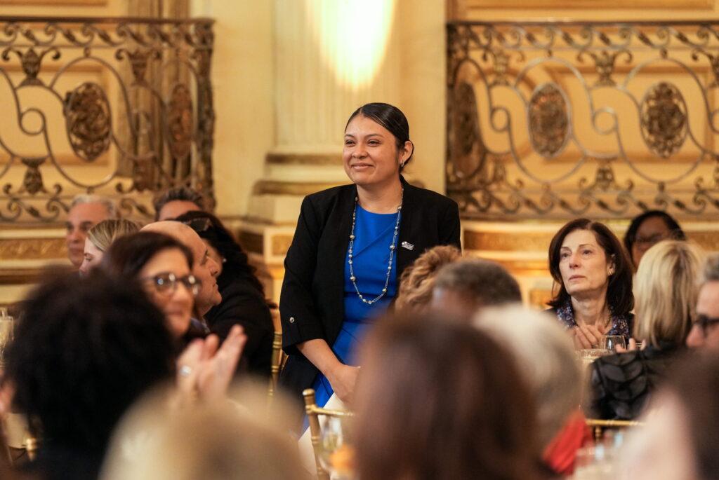 A person wearing a blue dress and black blazer is acknowledged in front of a crowd.