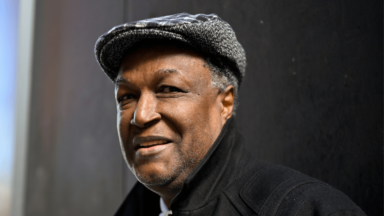 A person stands outdoors against a black wall, wearing a grey flat cap and black high-collared coat over a white shirt. Beyond the wall is a bright city street, buildings, and pedestrians blurred in the background.